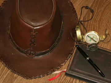 Brauner Hut, Kompass und Notizbuch auf Holzoberfläche.