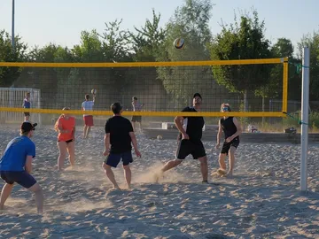 Sandplatz, Volleyballspieler springen, sonniger Tag, Bäume im Hintergrund.