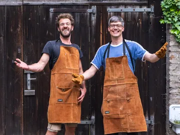 Two men in leather aprons gesture happily in front of the door.