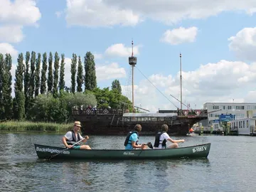 Three people paddle in a canoe in front of an old ship.