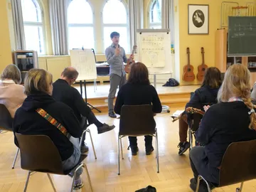 Mann spricht mit Gitarre zu sitzendem Publikum im Raum.