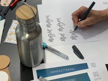 Metal bottle stands on table while hand lettering is practiced.