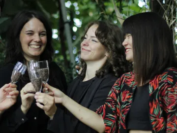 Three women with glasses make a toast in a green setting.