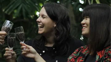Two women with glasses make a toast in a green setting.