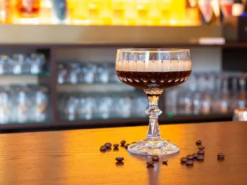 Cocktail glass with dark drink and coffee beans on table.