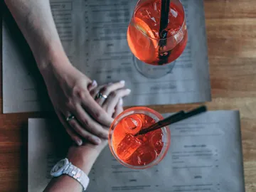 Two cocktails are sitting on a table, hands touching each other.