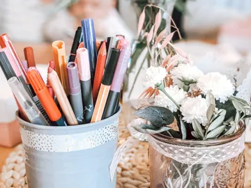 Colourful pens are placed in a container next to a flower vase.