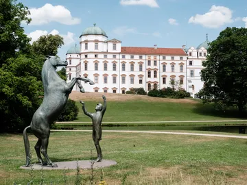 Eine Skulptur zeigt ein Pferd, das auf den Hinterbeinen steht, während ein Kind es anblickt. Im Hintergrund ist ein großes, historisches Gebäude und ein Park mit Bäumen.