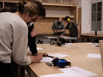 Two people are sketching at a table in a workshop.