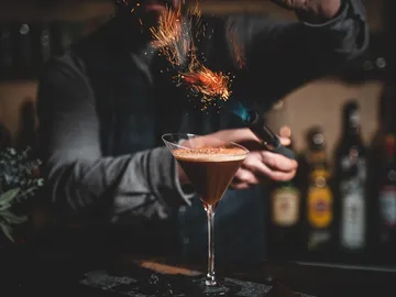 Cocktailglas mit Funken, während ein Barkeeper flambiert.