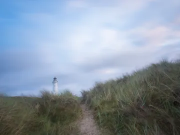 Lighthouse stands between grass dunes under a cloudy sky.