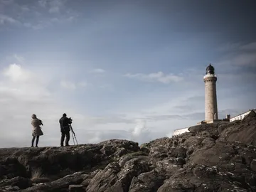 Two photographers are standing on rocks, watching a lighthouse.