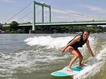 Surferin reitet eine Welle unter einer Brücke an einem Fluss.