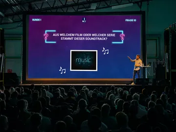A presenter asks questions about a musical soundtrack on stage.
