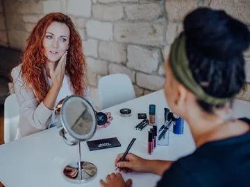 Woman looks at herself in the mirror while others apply makeup.