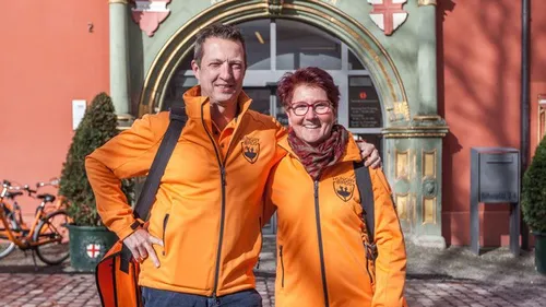 Zwei lächelnde Menschen in orangefarbenen Jacken, die draußen stehen.