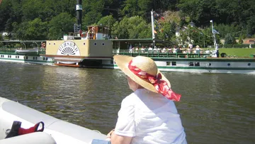 Frau mit Strohut betrachtet Dampfschiff auf einem Fluss.
