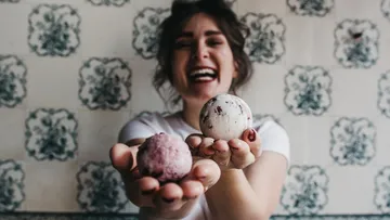 Frau hält zwei Eisballen vor dekorativem Hintergrund und lächelt.