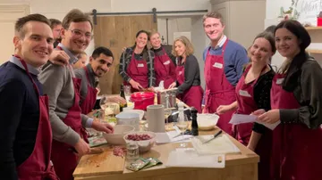 Group cooks together in a kitchen, everyone is wearing aprons.