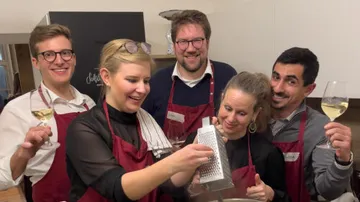 Fünf Personen in Schürzen kochen und lachen zusammen.