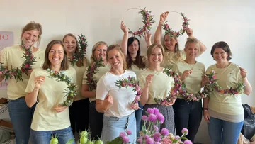Ten women hold flower-decorated wreaths in a room.