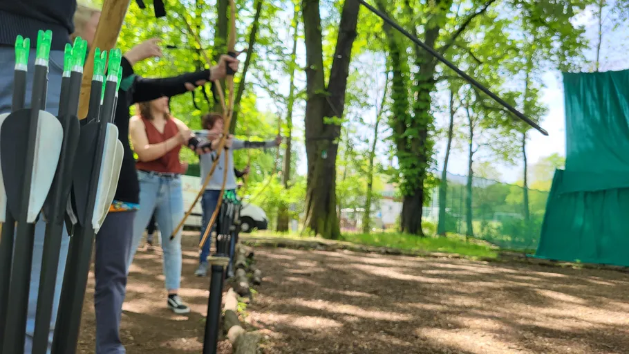 Pfeile stehen bereit; Personen zielen mit Bögen im Freien.