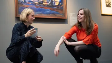 Zwei Frauen unterhalten sich neben einem Gemälde in einer Galerie.