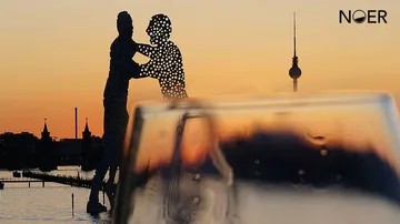 Skulpturen umarmen sich vor einem orangefarbenen Sonnenuntergang.