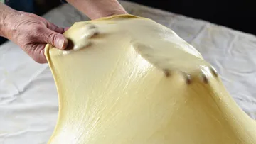 Hände dehnen einen gelben, elastischen Teig auf einem Tisch.