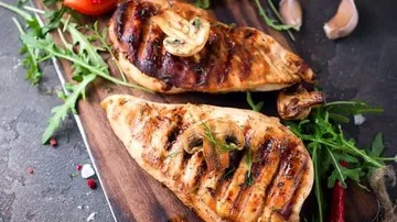 Gegrilltes Hähnchenbrustfilet liegt auf Holzbrett, umgeben von Gemüse.