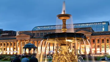 Brunnen sprudelt Wasser, umgeben von Gebäuden und Menschen.