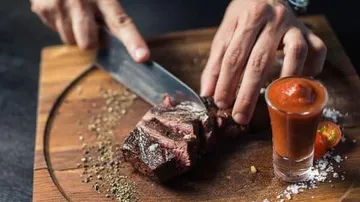 A steak is cut on a wooden board with sauce next to it.