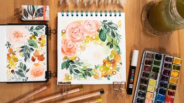 Skizzenblock mit Aquarellblumen, umgeben von Kunstmaterialien.