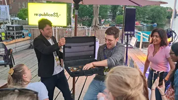 Zwei Männer öffnen einen Koffer vor Publikum am Fluss.