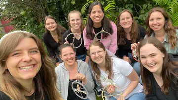 Neun Frauen halten Handarbeiten in einem Garten und lächeln.