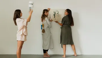 Drei Frauen hängen Kunstwerk an eine weiße Wand.