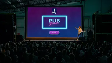 Ein Bildschirm zeigt „PUB quiz“, jemand erklärt. Publikum schaut zu.