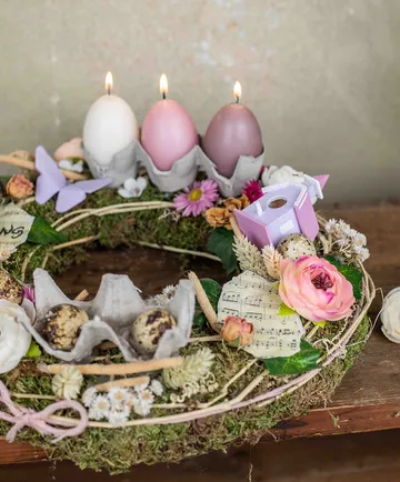 Osterkranz mit Kerzen, Blumen und bunten Eiern auf Tisch.