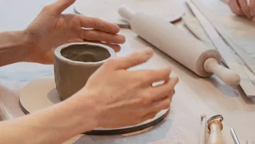 Hands form an unfired clay pot on a table.