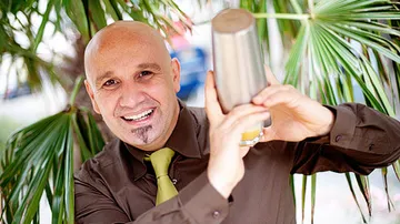Bartender shakes cocktails against a palm tree background, smiles friendly.