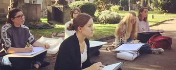 Five women are drawing on sketchpads while sitting outdoors.
