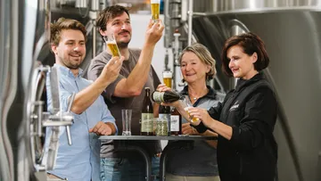 Vier Personen stoßen mit Bier im Brauerei-Umfeld an.