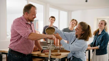 Eine Frau gibt Körner in eine Mühle, Leute beobachten.