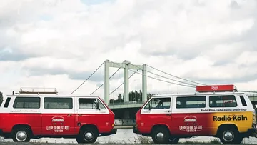 Two red vans are standing in front of a bridge by the river.
