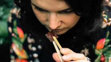Woman holding food with chopsticks, intensely concentrated, green environment.