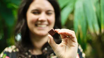 Woman holding a piece of chocolate in a tropical setting.