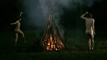 Zwei nackte Personen tanzen um ein großes Feuer.