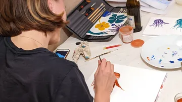 Artist paints on paper, surrounded by painting supplies.