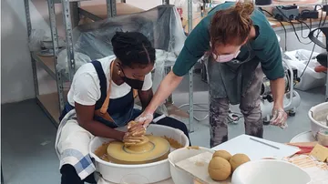 Two women make clay on a potter's wheel in a studio.