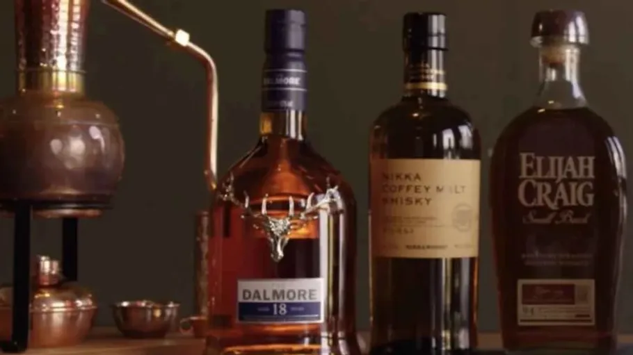 Three whiskey bottles are standing on wood next to a still.